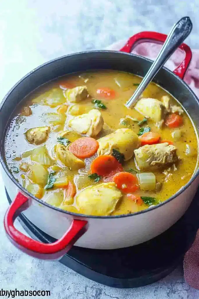 A pot of Jamaican chicken soup with tender chicken, carrots, and potatoes in a flavorful golden broth, served with a spoon.