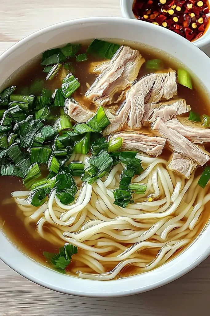 A vibrant bowl of noodle soup topped with herbs, chili flakes, and tender meat slices.